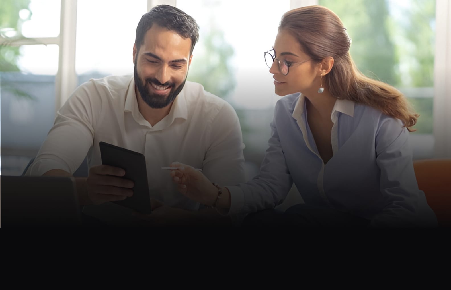 Two professionals collaborating, with the man smiling while reviewing a partnership agreement on a device and the partner attentively observing.